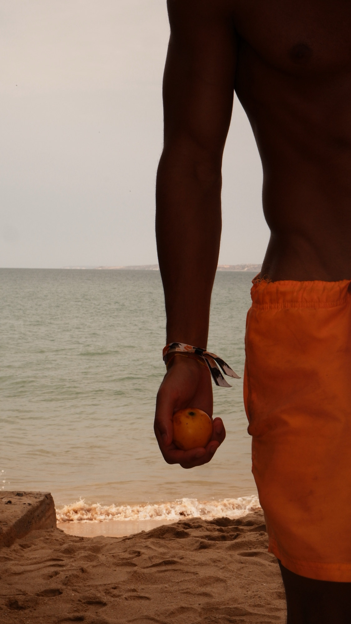 Man in orange shorts holding a fruit by the sea