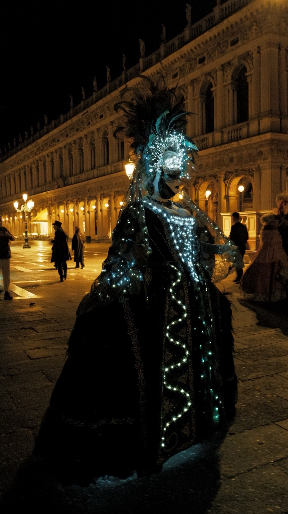 A masked figure in a gown lit with small lights in a night square